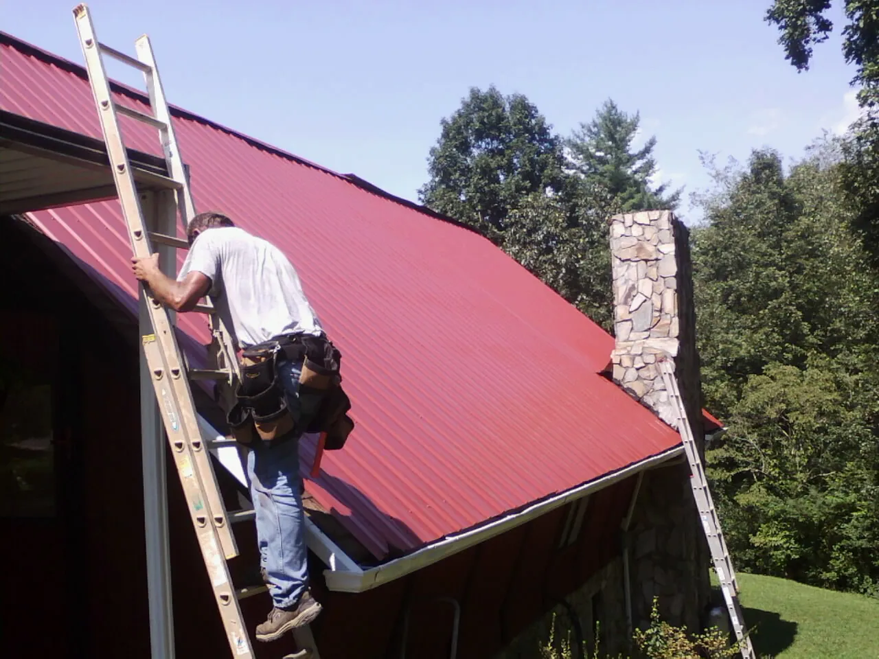 Roofer on a ladder beside a red metal roof and stone chimney — Soffit & Fascia Repair by Roofing Brothers in Gridley